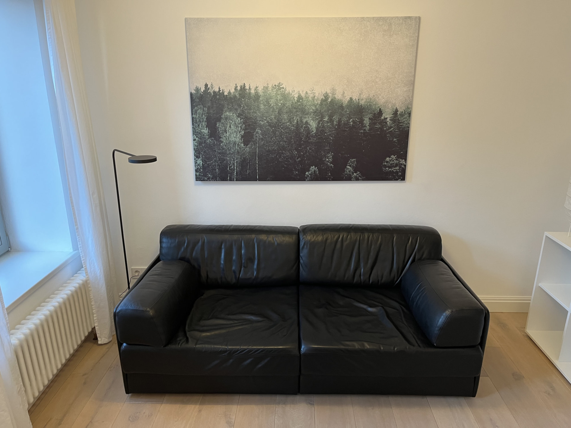 Helles Wohnzimmer mit schwarzem Ledersofa und Waldbild in einer möblierten Wohnung auf Zeit